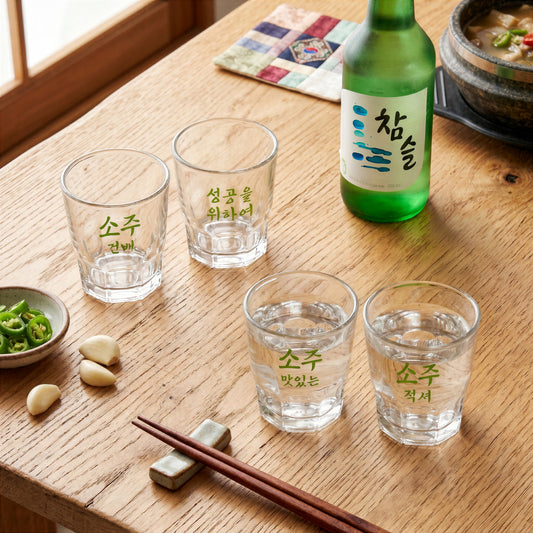 A lifestyle product photo of the Final Touch Soju Glasses Set of 4, featuring clear glass Korean-style shot glasses with unique green Hangul text on a wooden table setup next to a traditional green soju bottle and chopsticks. Two of the four multi-faceted heavy-base shot glasses are filled with clear soju.