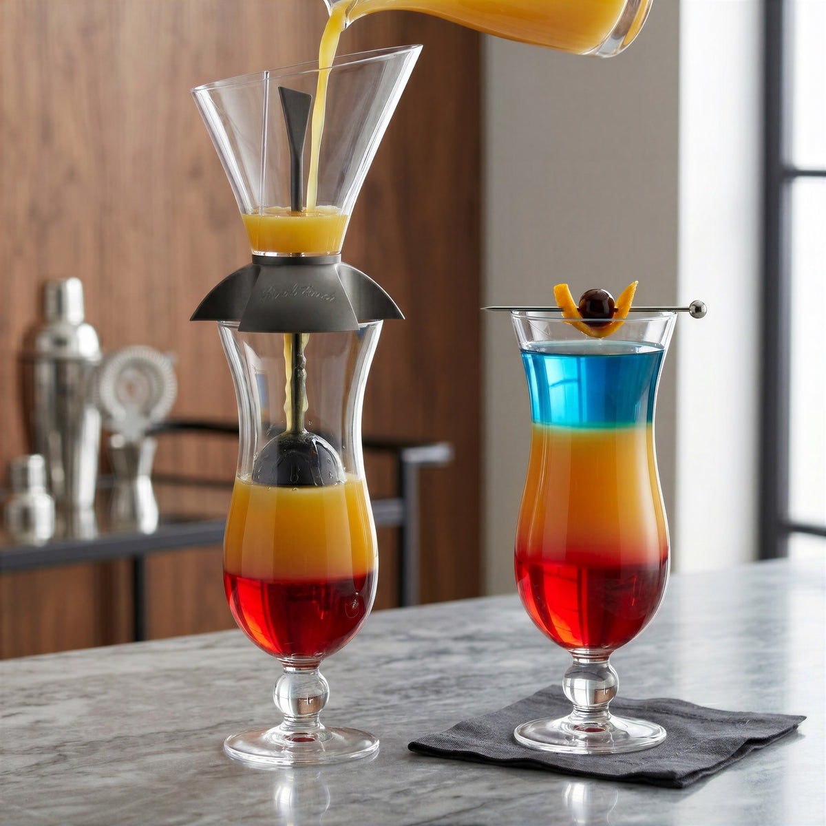 Pouring a layered drink using the Final Touch cocktail layering tool on a hurricane glass with a floating black ball, next to a completed red, orange, and blue layered cocktail on a marble bar.