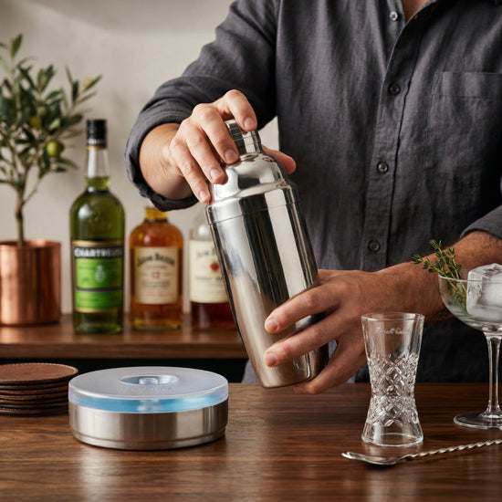 Person shakes stainless steel cocktail shaker on wooden bar with bottles and glasses in background.