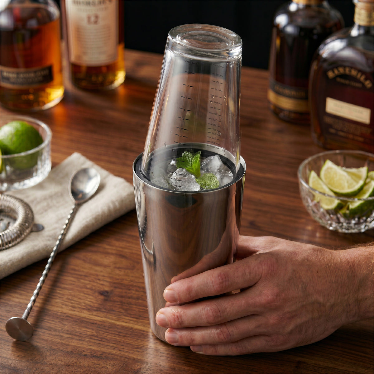 Final Touch Double-Wall Boston Shaker with a stainless steel cocktail shaker tin and a clear glass measuring beaker insert, shown on a wooden home bar with fresh cocktail ingredients.
