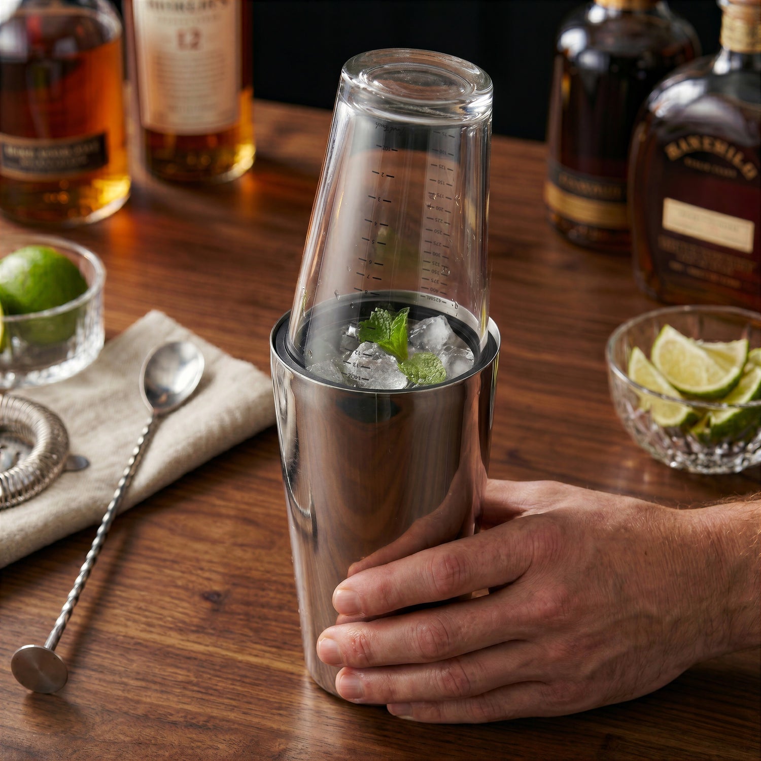 Final Touch Double-Wall Boston Shaker with a stainless steel cocktail shaker tin and a clear glass measuring beaker insert, shown on a wooden home bar with fresh cocktail ingredients.