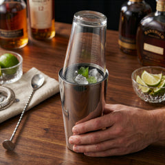 Final Touch Double-Wall Boston Shaker with a stainless steel cocktail shaker tin and a clear glass measuring beaker insert, shown on a wooden home bar with fresh cocktail ingredients.