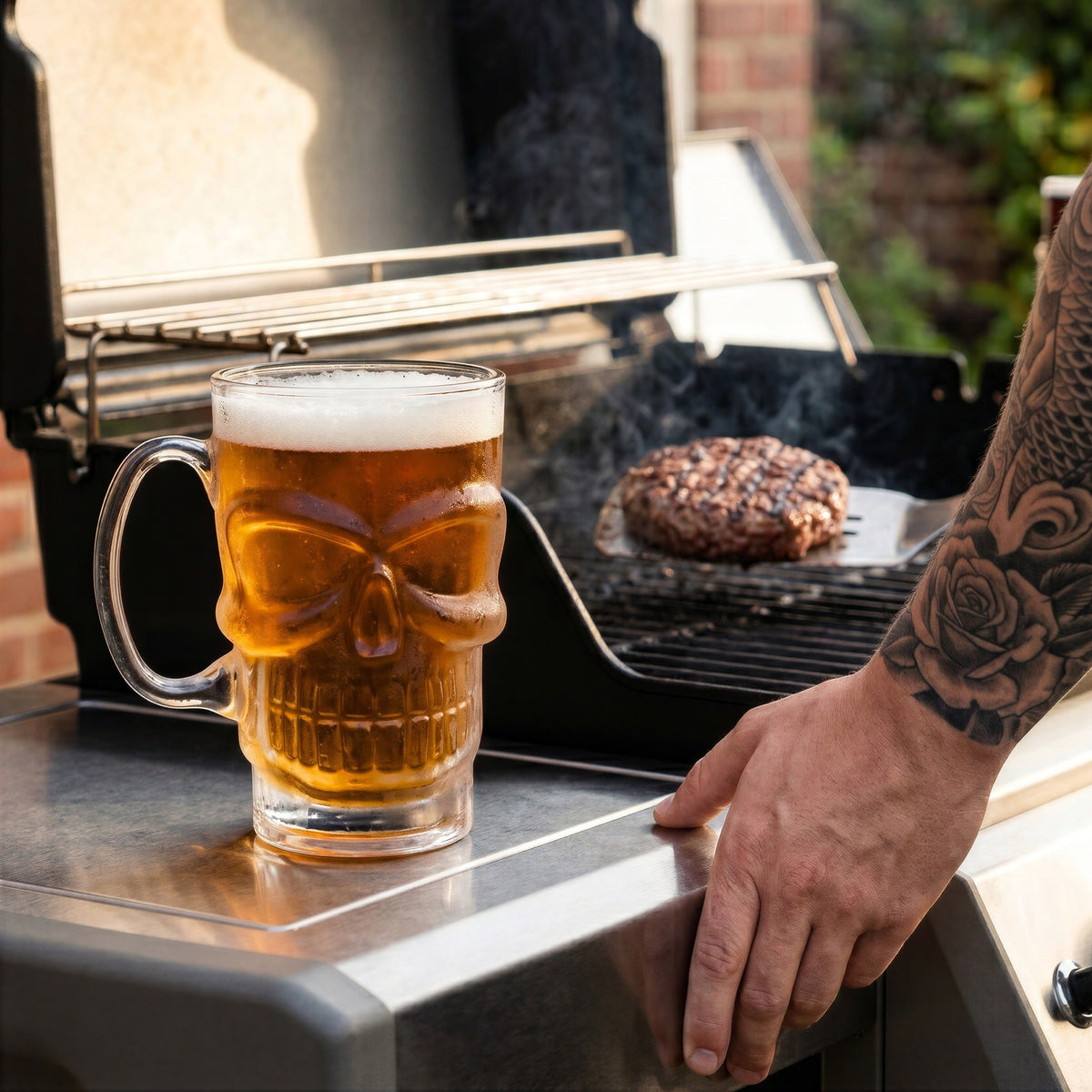 A close-up lifestyle photo of the Final Touch Brain Freeze Skull Beer Mug filled with amber beer and foam, sitting on the side shelf of a clean stainless steel barbecue grill while a person with a full sleeve tattoo uses a spatula to flip a burger on the grates.