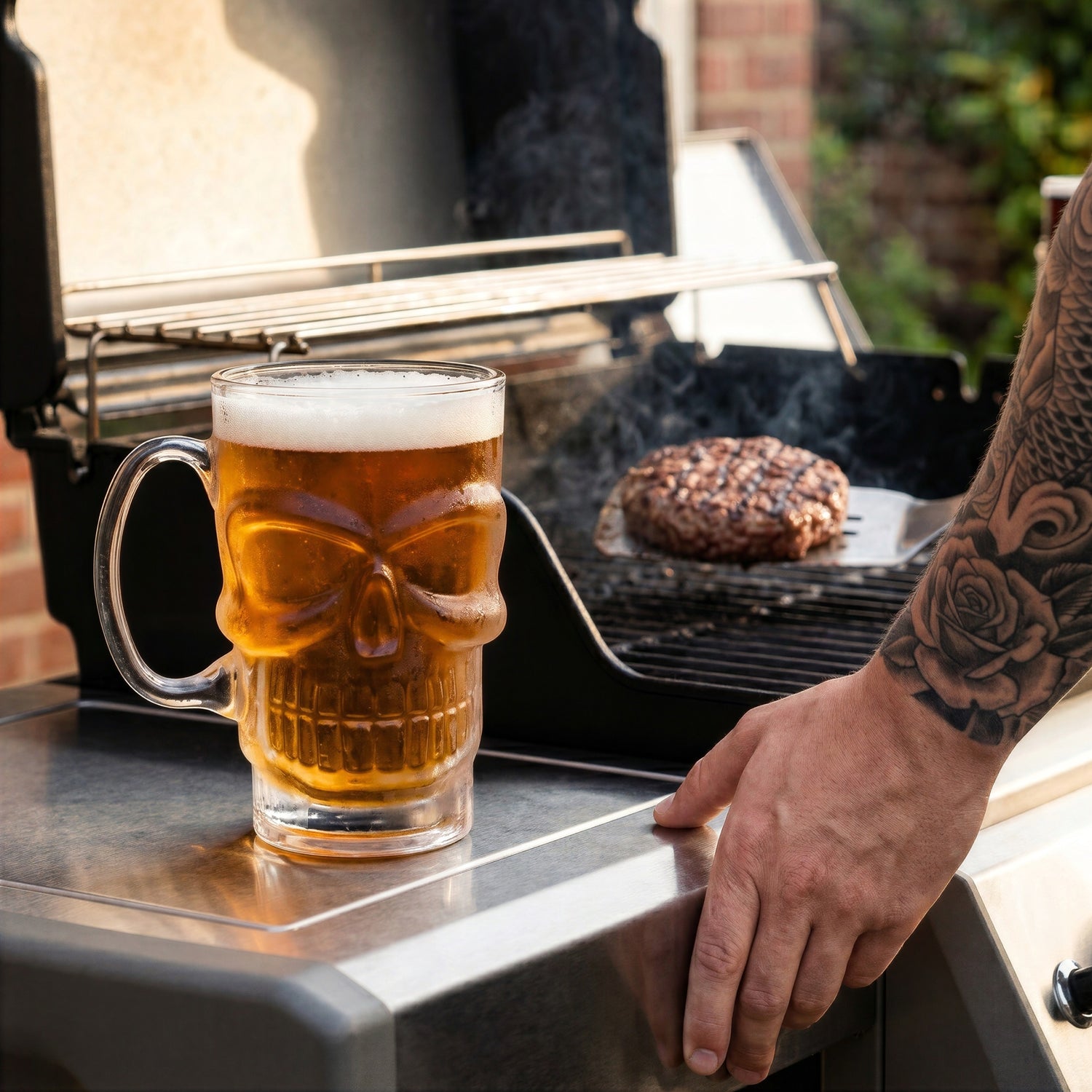 A close-up lifestyle photo of the Final Touch Brain Freeze Skull Beer Mug filled with amber beer and foam, sitting on the side shelf of a clean stainless steel barbecue grill while a person with a full sleeve tattoo uses a spatula to flip a burger on the grates.