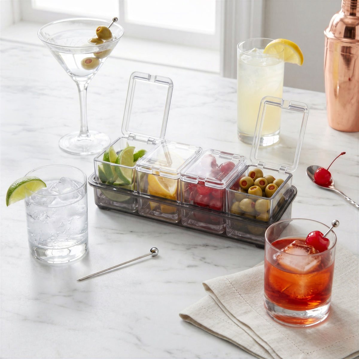 Final Touch 5 Piece Garnish Bar Caddy on a marble counter next to various cocktails including a martini and an old fashioned. The clear 4-compartment acrylic organizer has individual hinged lids open to show fresh lime, lemon, cherries, and olives.