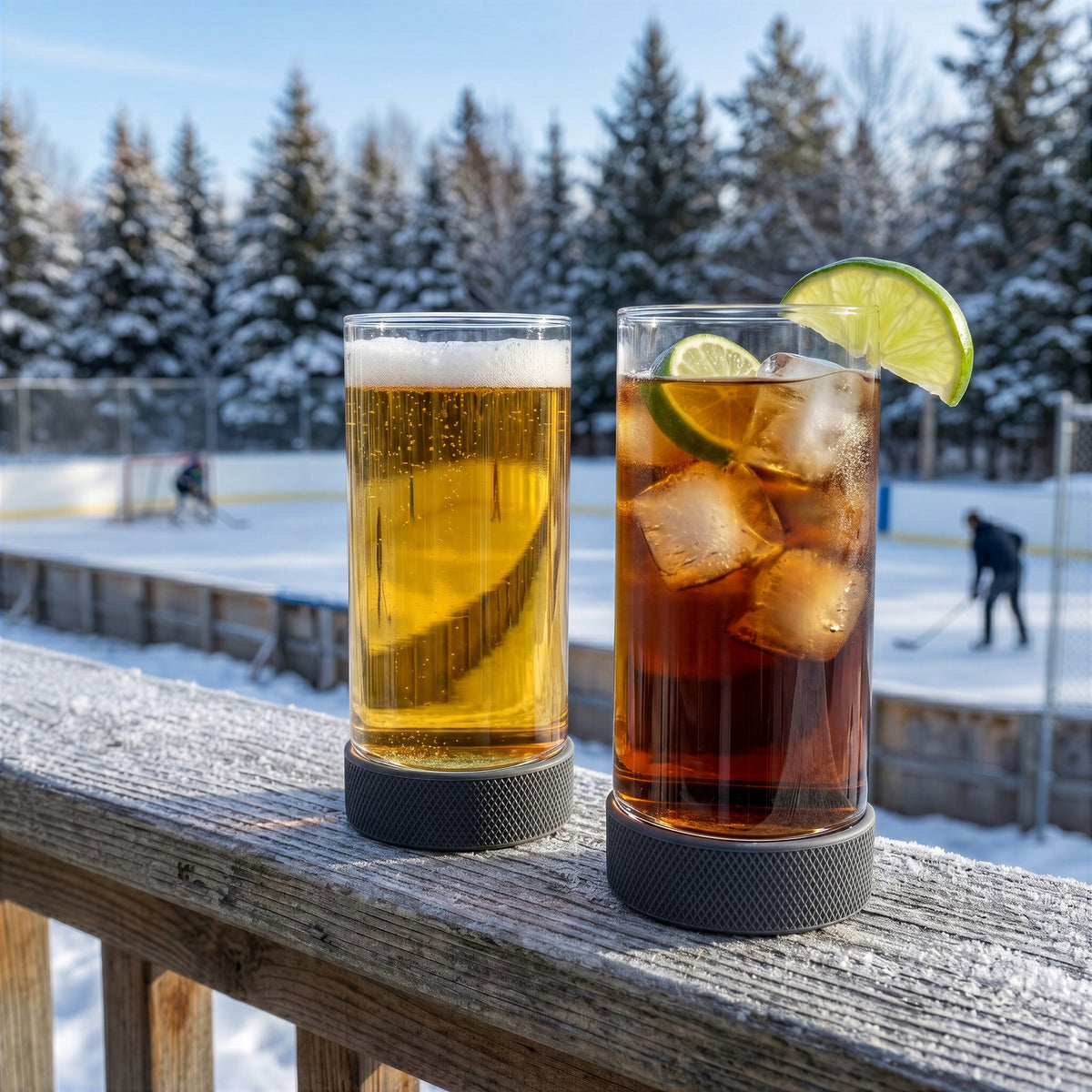 Two Final Touch Breakaway Hockey Puck Pint glasses filled with beer and a mixed drink, sitting on a snowy winter deck overlooking an outdoor backyard hockey rink surrounded by pine trees.