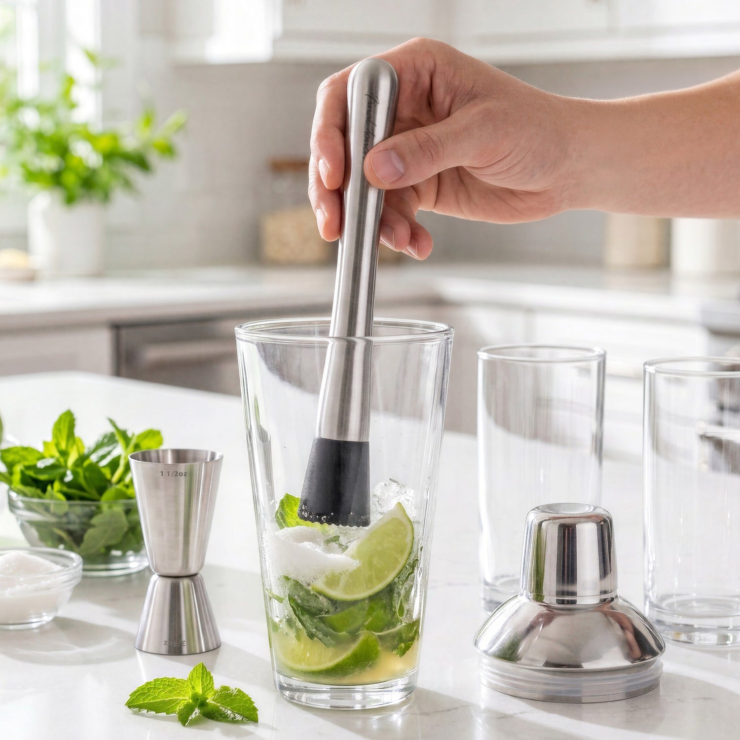 Final Touch stainless steel cocktail muddler with a black head being used to muddle fresh mint, lime, and sugar in a glass alongside a matching jigger and shaker lid on a bright kitchen counter