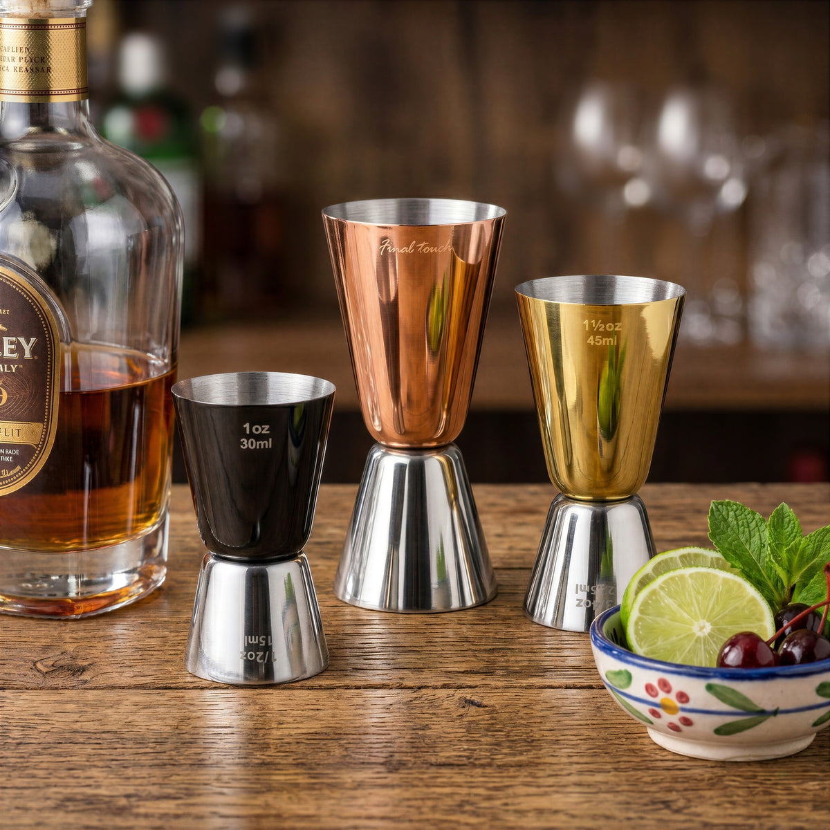 Three double-sided cocktail measuring cups from the Final Touch Mixed Metal Jigger Set in black, copper, and gold finishes on a rustic wooden bar counter.