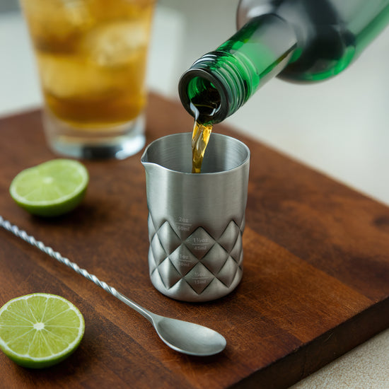 Stainless steel jigger with diamond pattern pours liquid onto wooden board near lime slices.