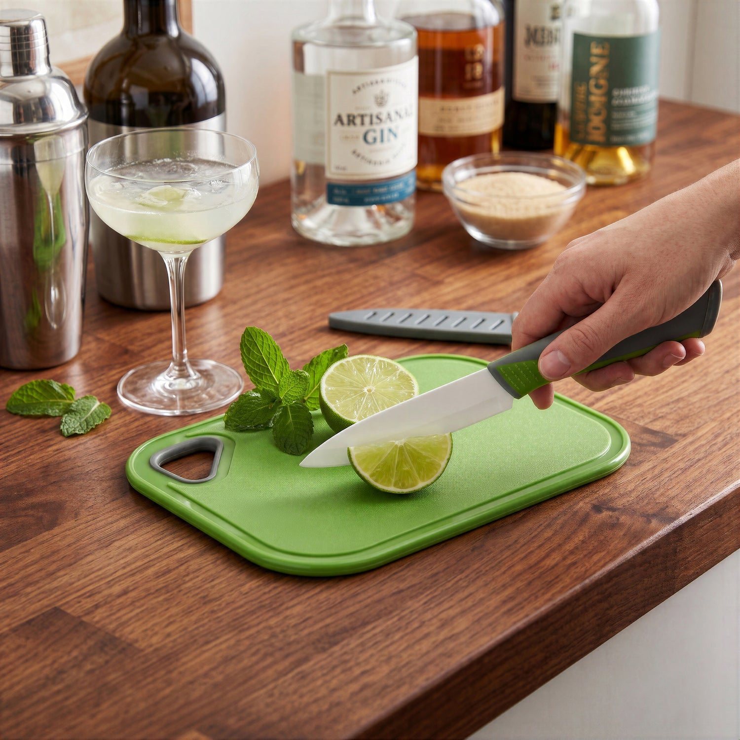 A close-up of a person slicing a fresh lime with a ceramic knife on a Final Touch non-slip bar cutting board. The green and grey handled ceramic knife is making a precise cut. In the background, a cocktail, shaker, and several liquor bottles like Artisanal Gin, whiskey, and brown sugar are on a dark wood surface.
