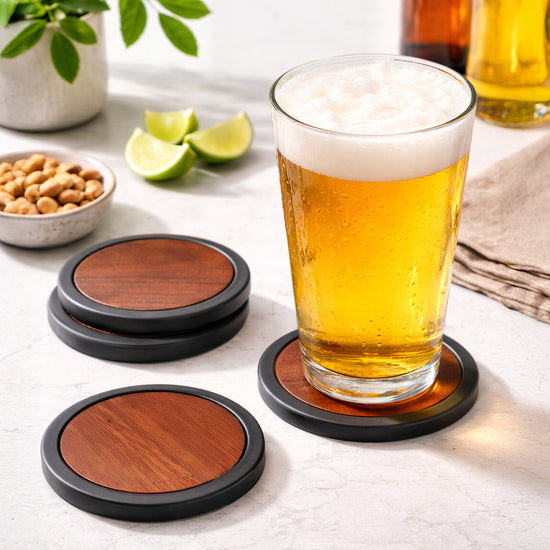 Set of four wooden coasters with black silicone edges, resting on a light surface near a glass of beer.