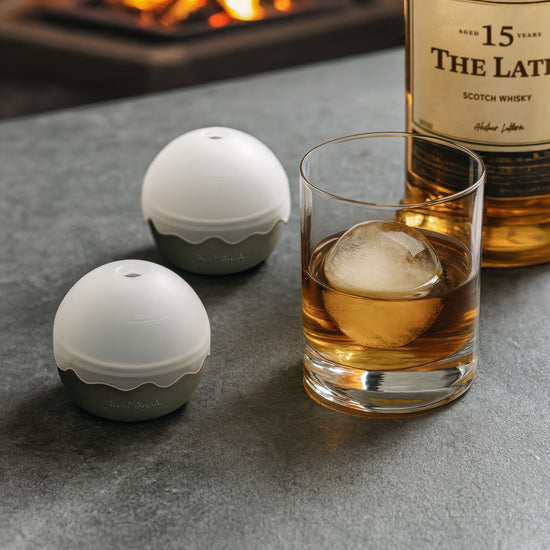 Two white and gray silicone ice ball molds on a stone table beside a glass of whiskey.