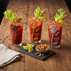 Set of 3 Final Touch Caesar Time cocktail glasses in black, gold, and silver graphics, filled with garnished Caesar cocktails on a rustic wooden bar table.