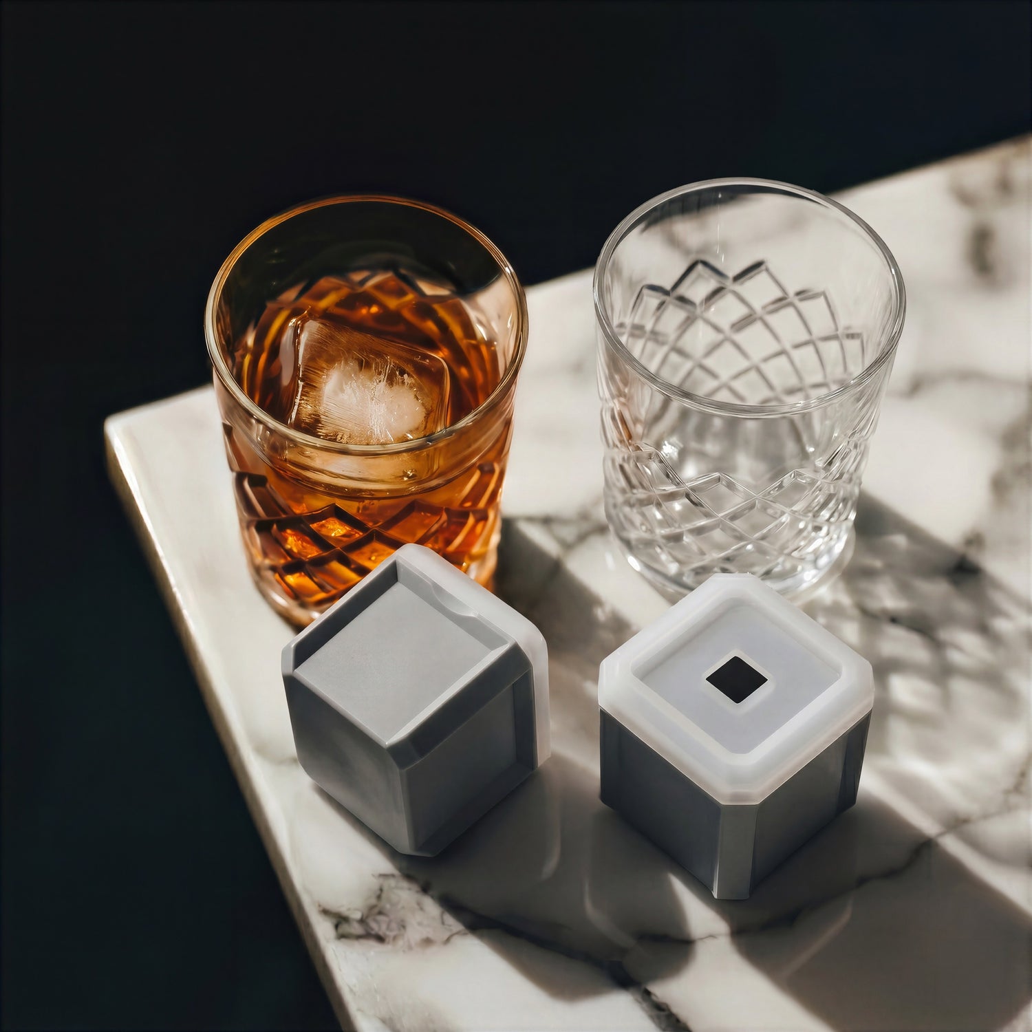 Final Touch Yarai Double Old-Fashioned Glass Set on a marble counter, featuring one crystal whiskey glass with a large clear square ice cube and grey silicone ice cube moulds.