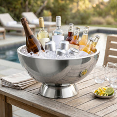 Final Touch Double-Wall Stainless Steel Beverage Bin with pedestal base, filled with ice cubes, unbranded champagne, white wine, vodka, cider bottles, and aluminum cans. The sleek, condensation-free metallic ice bucket sits on a rustic wooden outdoor table by a backyard swimming pool, styled with cocktail glasses and fresh lime garnishes.
