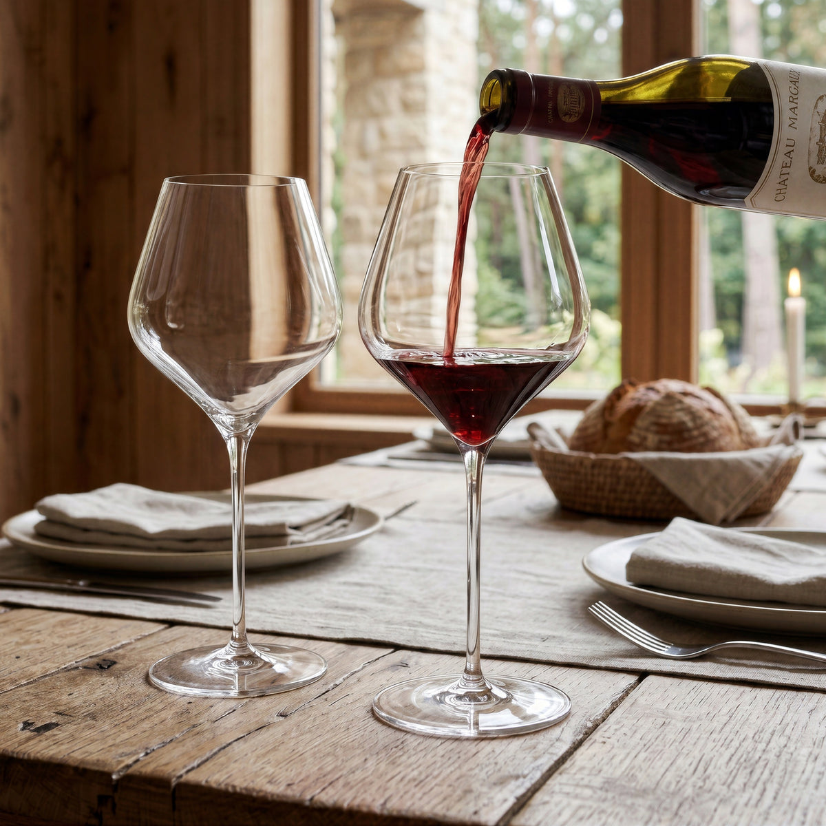Pair of large, elegant Final Touch Burgundy crystal wine glasses with thin rims on an upscale rustic cottage dinner table, with rich red wine being poured from a bottle into one glass.
