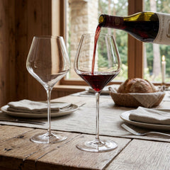 Pair of large, elegant Final Touch Burgundy crystal wine glasses with thin rims on an upscale rustic cottage dinner table, with rich red wine being poured from a bottle into one glass.