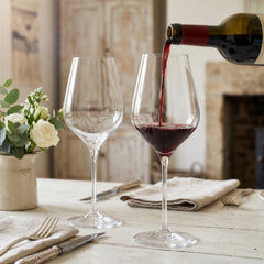 Pair of large, elegant Final Touch Bordeaux crystal wine glasses with thin rims on an upscale rustic cottage dinner table, with rich red wine being poured from a bottle into one glass.