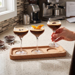 A hand lifting a coupe glass from the Final Touch Espresso Martini Flight Set, styled on a kitchen countertop next to an espresso machine and coffee beans.