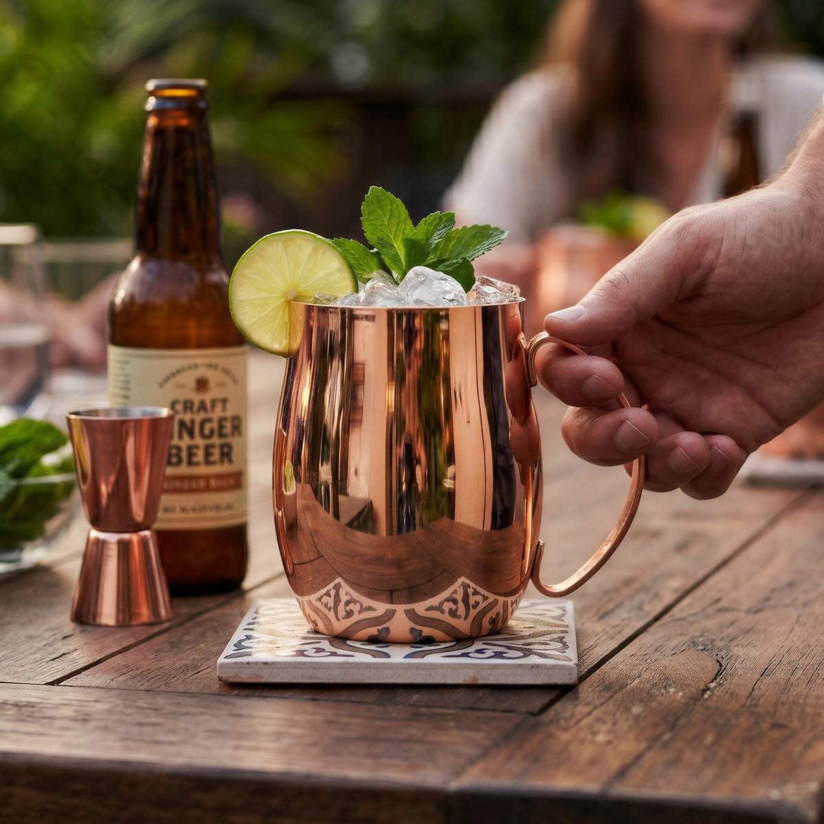 A highly polished copper Final Touch Double-Wall Mule Mug filled with a Moscow Mule cocktail, mint garnish, and lime wheel, held by a hand on a rustic wooden outdoor patio table during a backyard gathering.