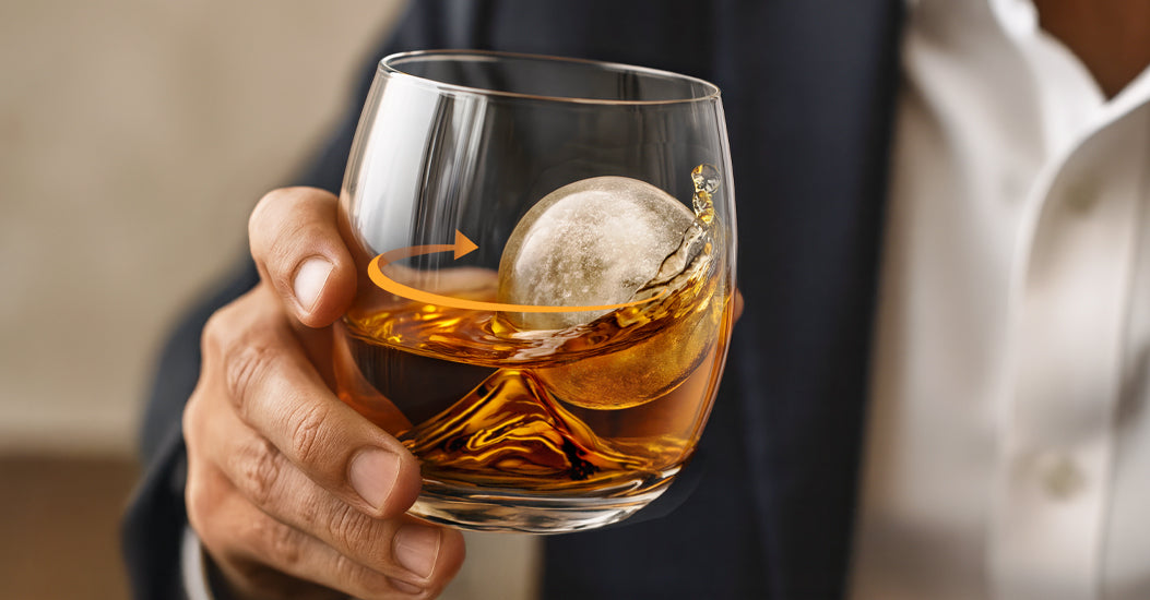 Man holds glass of amber liquid with large ice ball in On The Rock glass.
