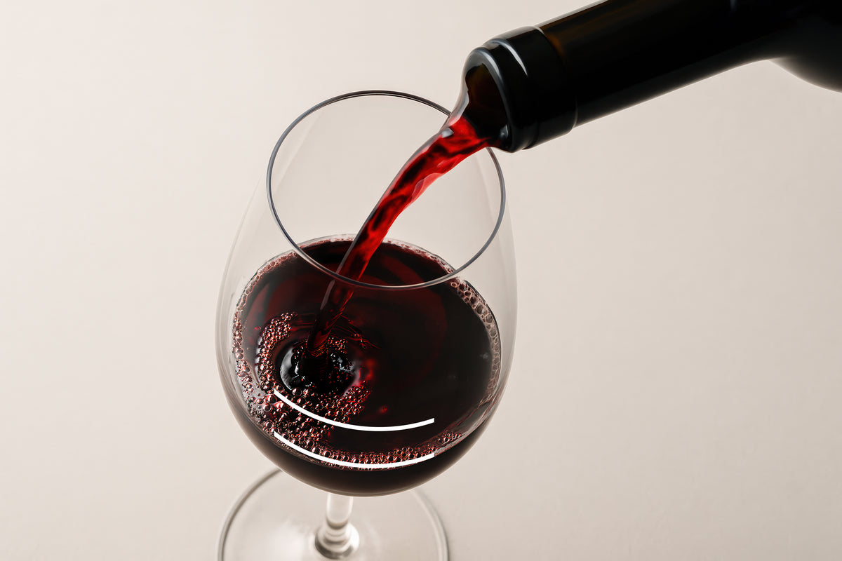 Red wine being poured from a dark bottle into a Final Touch Wine Tasting Glass against a light beige background.