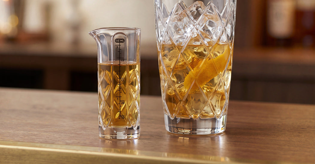Yarai-pattern glass measuring jigger beside a tall mixing glass with ice, an amber cocktail and citrus peel on a bar counter.