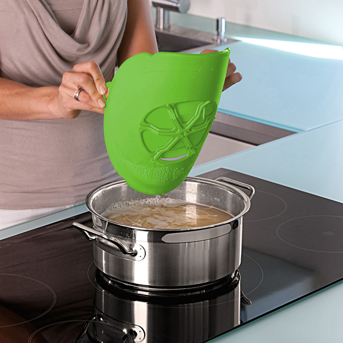 A person uses a green, silicone Boil Guard over a pot on the stove.