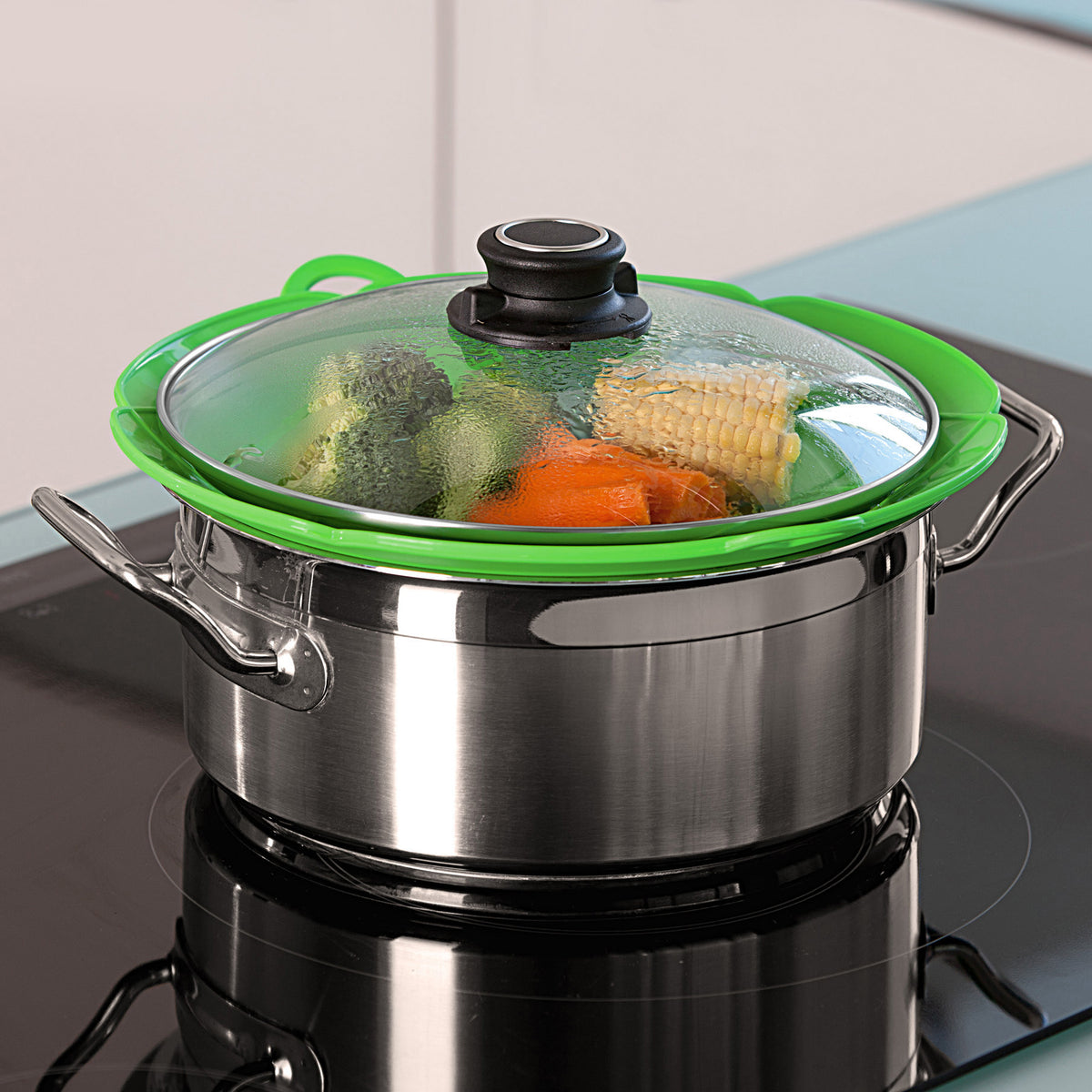 Green and clear lid fits over stainless steel saucepan with visible vegetables inside.
