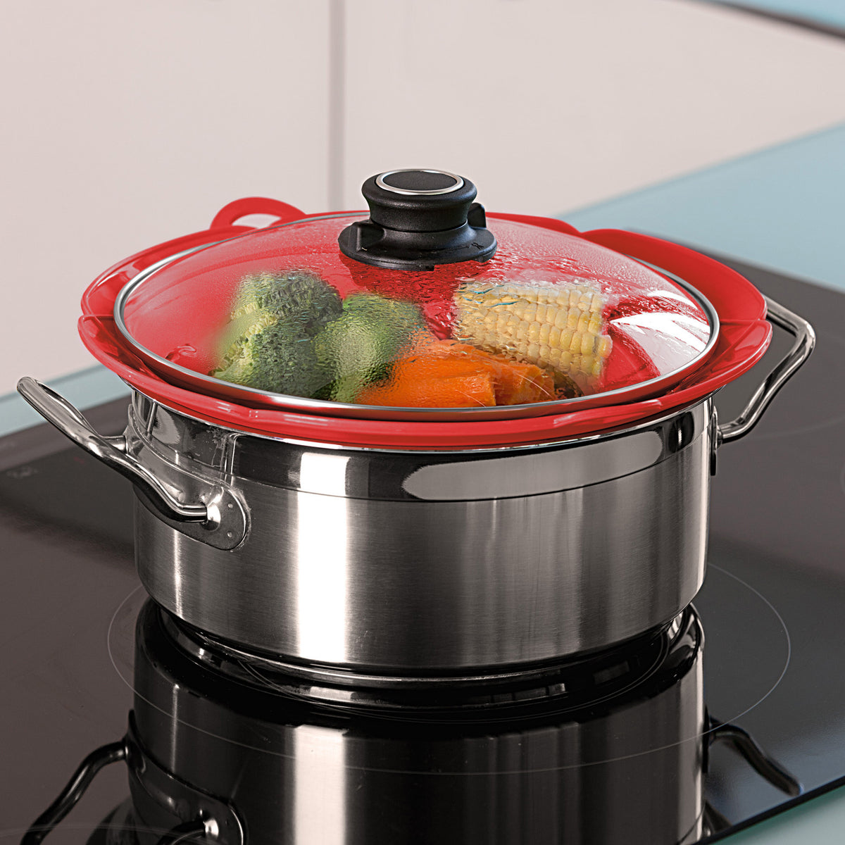 Red and silver saucepan with a transparent lid, filled with vegetables on a stovetop.