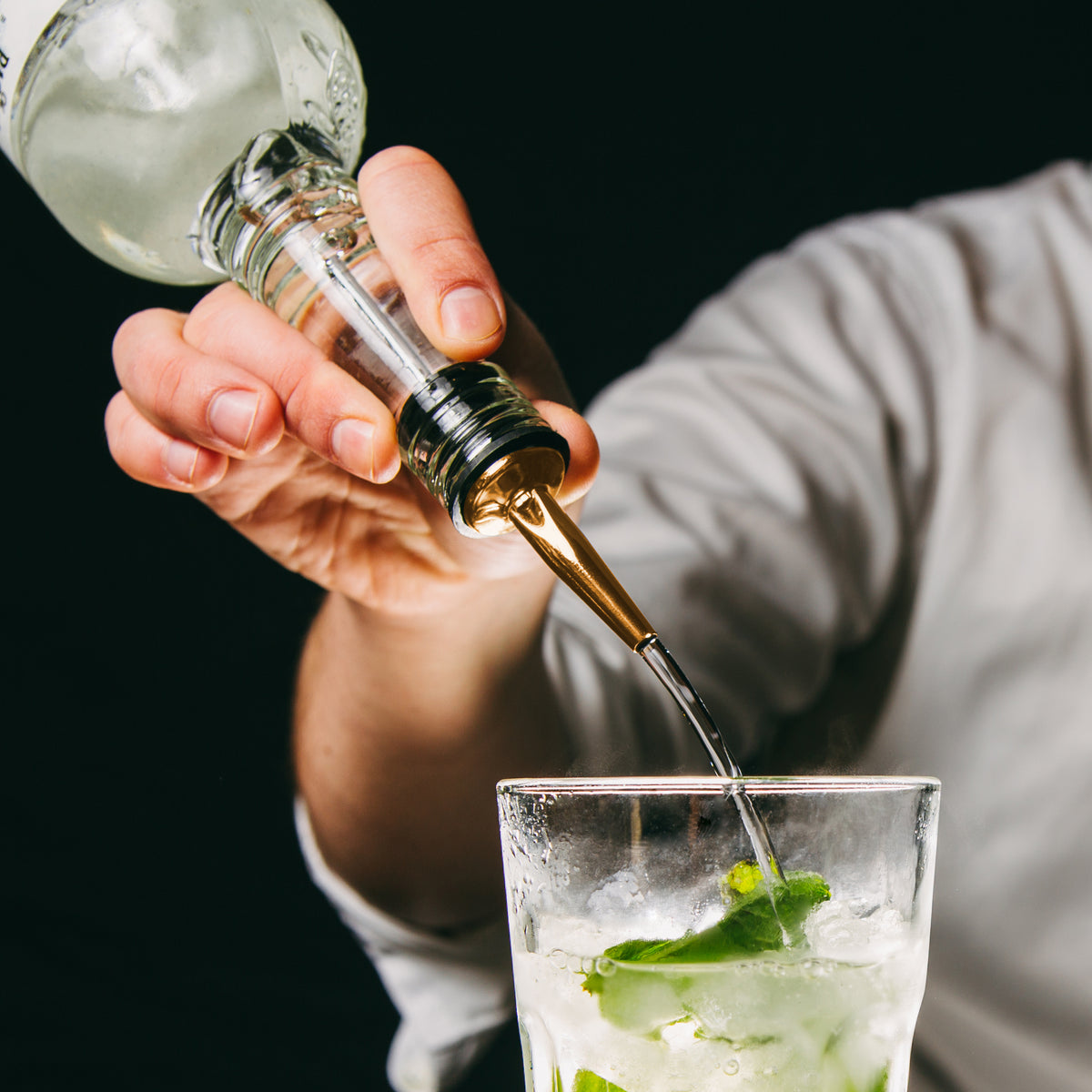 Hand pours clear liquid from stainless steel bottle pourer into glass with ice and mint.