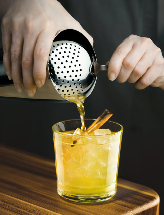 Stainless steel julep strainer pours golden liquid into a glass with ice and cinnamon.